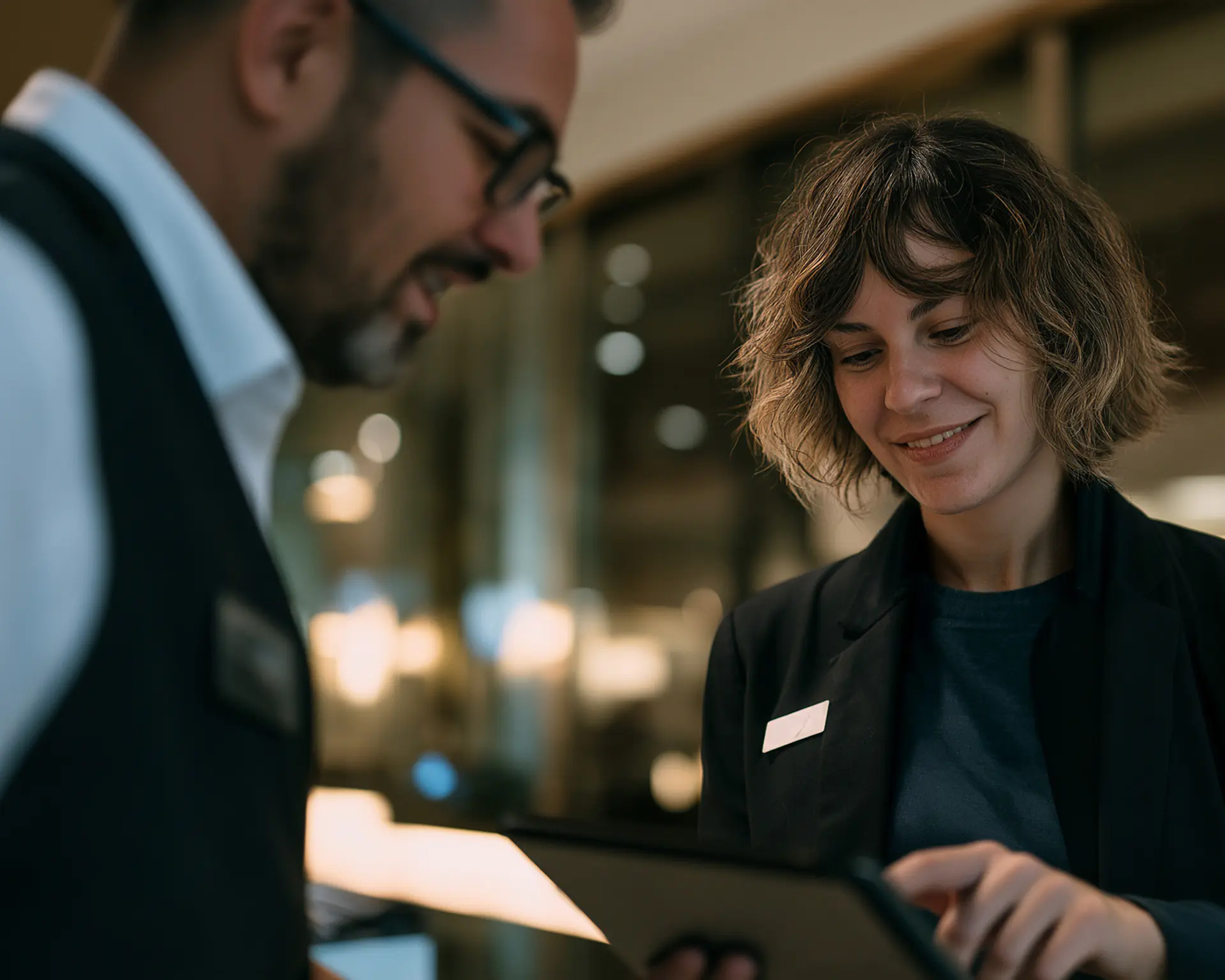 Two people in business attire stand indoors. One person holds a tablet and smiles whilst showing it to the other, who is slightly out of focus. Both wear name badges, suggesting a professional or service setting focused on hotel employee performance.