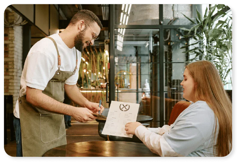A waiter wearing an apron takes an order from a seated woman holding a menu in a modern restaurant with plants and large windows in the background.