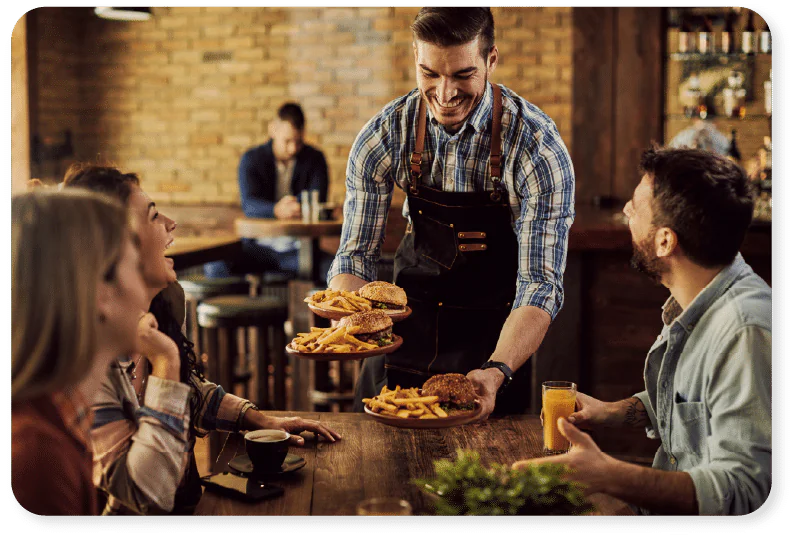 A smiling waiter delivers plates of burgers and chips to a group of three people sitting at a wooden table in a cosy restaurant. One person holds a drink, and everyone appears happy and engaged.