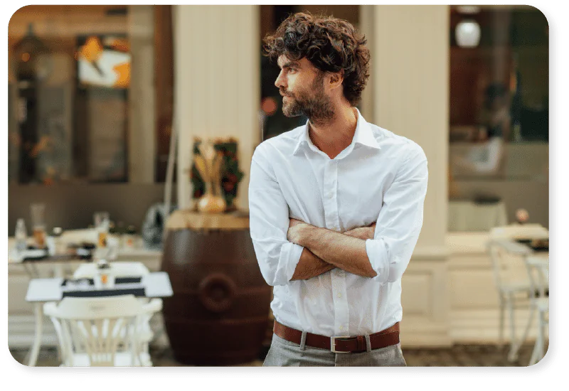 A man with curly hair and a beard, wearing a white shirt and brown belt, stands outside with arms folded, looking to the side. There are tables, chairs, and decorations in the blurred background.
