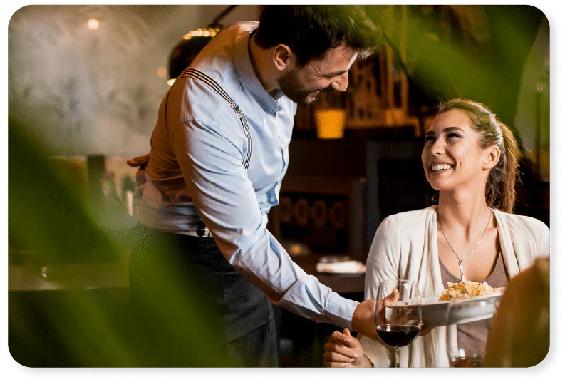 A smiling woman sits at a restaurant table with wine, receiving a plate of food from a cheerful waiter. Green leaves partially frame the scene.