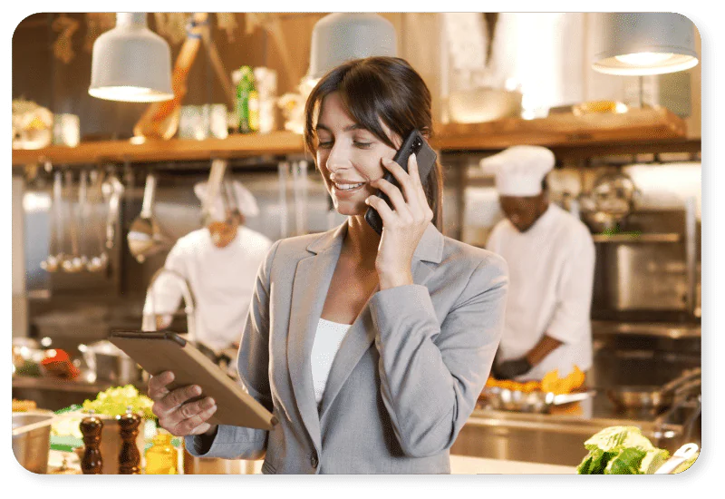 A woman in a suit smiles whilst talking on the phone and holding a tablet in a busy restaurant kitchen, with chefs working in the background.