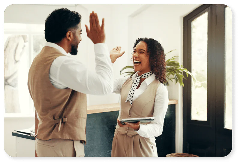 Two people in business attire laugh and share a high five in a bright office setting, with one holding a tablet and a blackboard and a potted plant in the background.