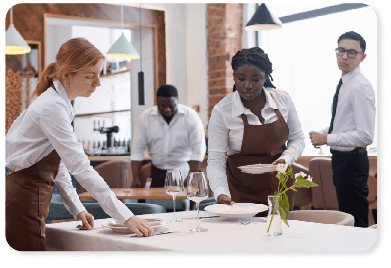 Four restaurant waiters in white shirts and brown aprons lay tables with plates and glasses in a modern dining area, preparing for service.