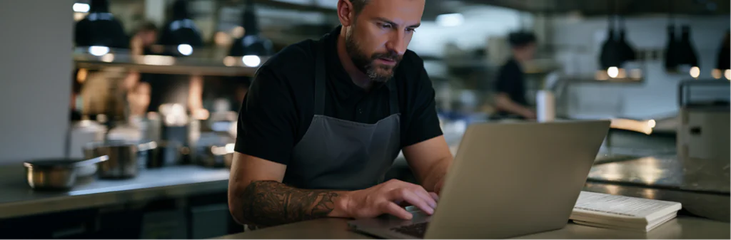 A man wearing an apron works on a laptop in a restaurant kitchen, with a notebook beside him and blurred kitchen staff in the background.