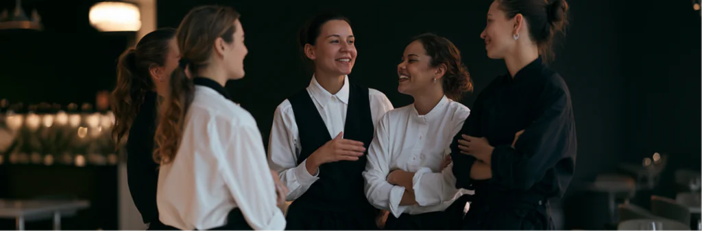 Five women in uniform stand together inside a restaurant, smiling and talking in a relaxed, cheerful manner. The background is softly lit and features tables and chairs, creating a warm, friendly atmosphere.