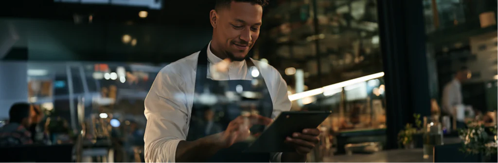 A smiling chef in a dark apron uses a tablet in a modern, well-lit restaurant, with shelves and diners visible in the background.