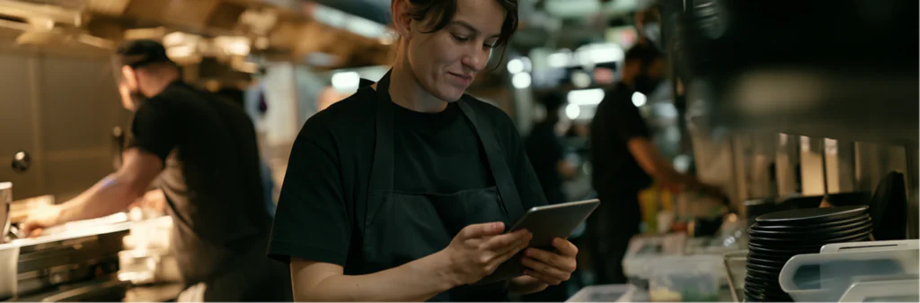 A person in a black apron smiles whilst using a tablet in a busy restaurant kitchen, with other staff working in the background.