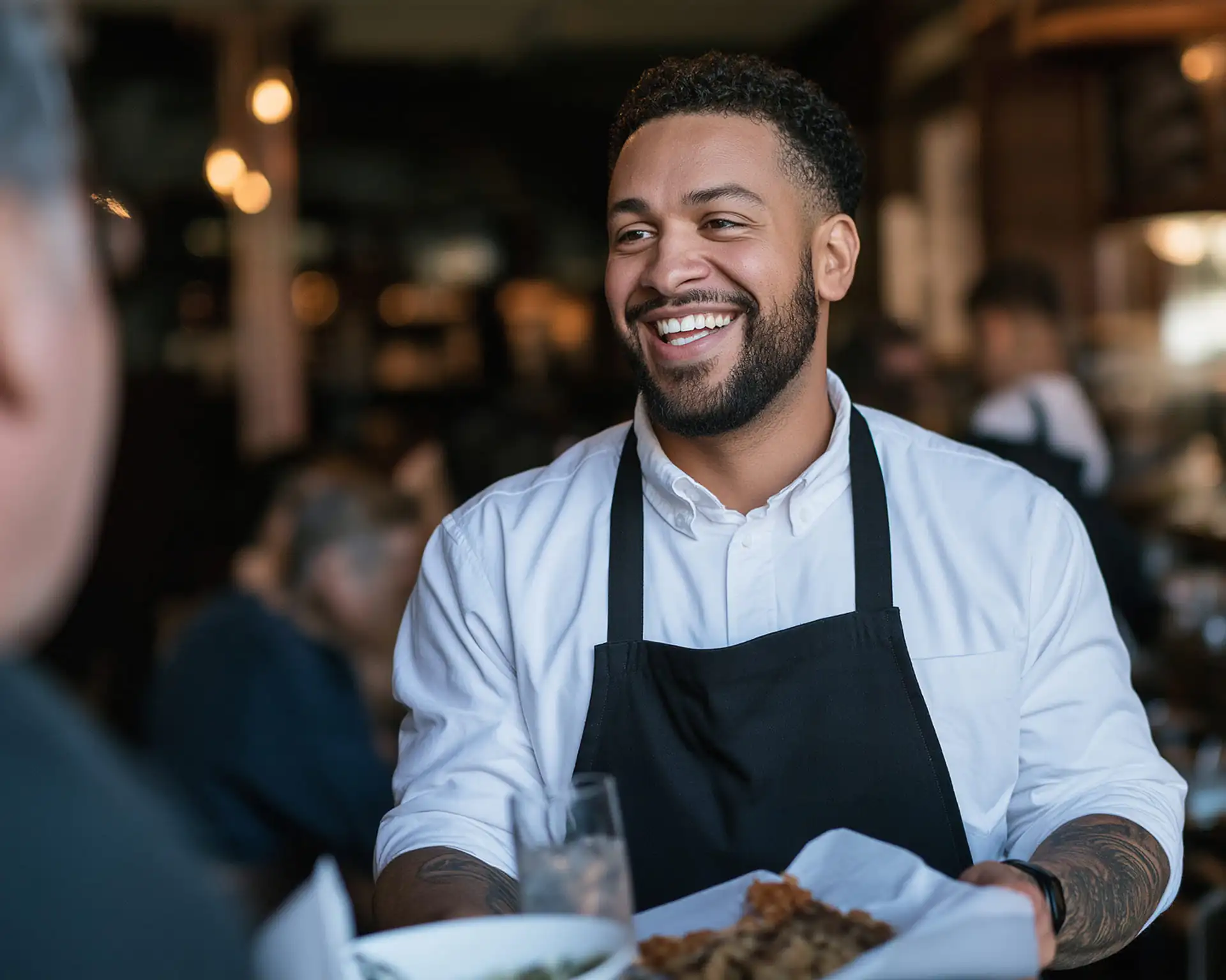 A smiling waiter in a white shirt and black apron holds a tray of food whilst speaking to a customer.