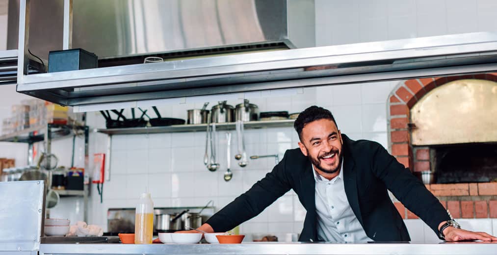 Un hombre vestido con traje sonríe y se asoma por encima de la encimera de una cocina moderna y profesional, con utensilios y un horno de ladrillo al fondo.
