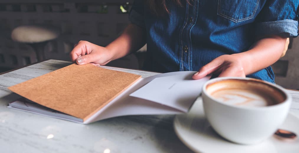 Una persona con una camisa vaquera lee un cuaderno abierto en una mesa, junto a una taza de café, en un acogedor ambiente interior.