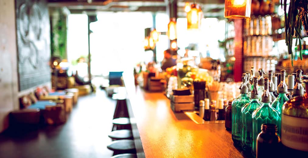Interior de un bar de moda con una larga barra de madera, diversas botellas y vasos, lámparas colgantes y una zona de asientos difuminada; la luz natural entra a raudales por los grandes ventanales.