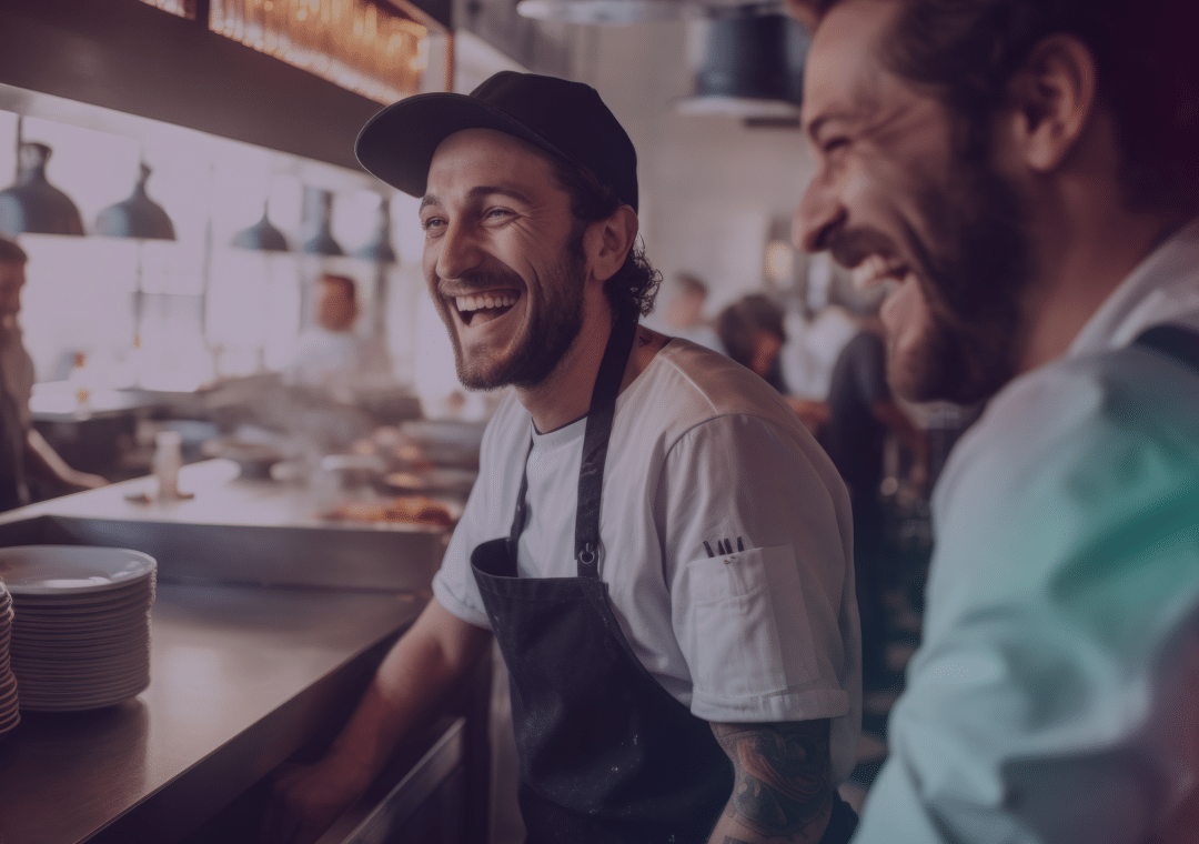 Dos cocineros, con delantales y gorros, se ríen juntos en la ajetreada cocina de un restaurante, rodeados de pilas de platos y una iluminación cálida, lo que crea un ambiente animado y acogedor.