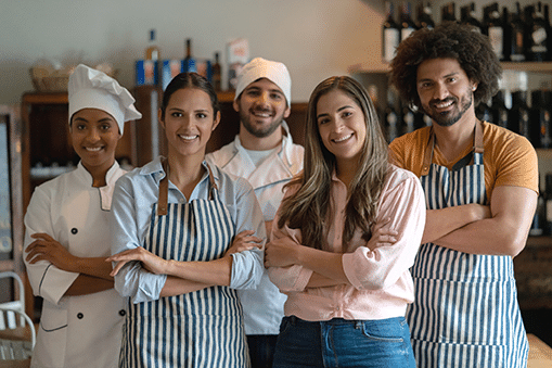 Cinco empleados de un restaurante, algunos con delantal y gorro de cocinero, posan juntos sonriendo con los brazos cruzados en el interior de un restaurante, con botellas de vino visibles al fondo.