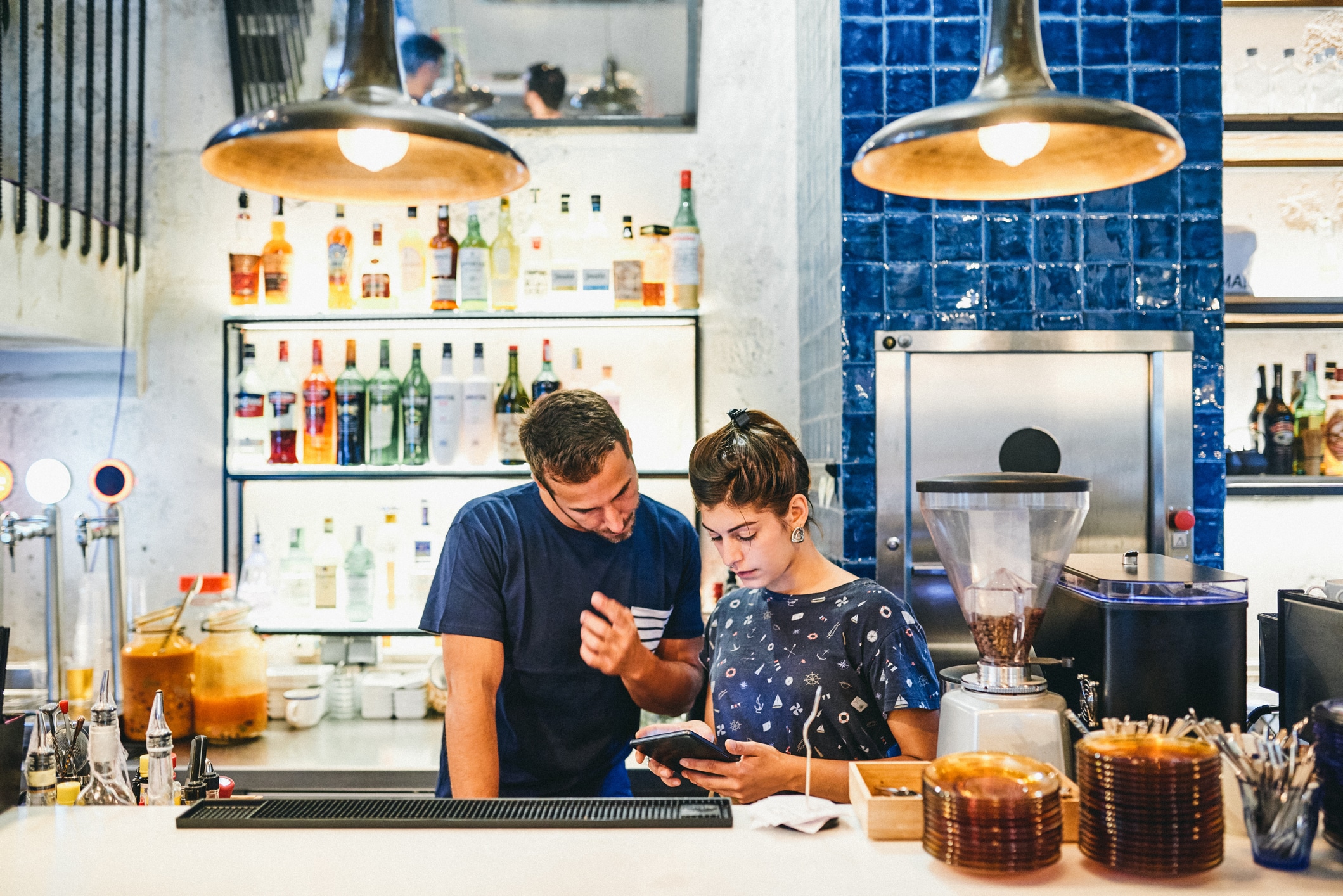 Dos personas están de pie detrás de la barra de un bar, hablando de algo en una tableta digital. Detrás de ellas se ven botellas de alcohol expuestas en las estanterías, y unas lámparas colgantes iluminan la escena. El ambiente parece informal y de concentración.