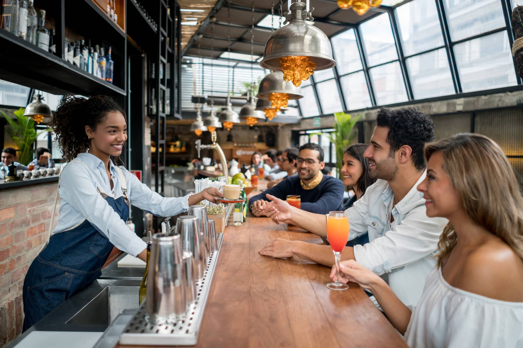 Un camarero sonriente sirve bebidas a un grupo de jóvenes sentados en una barra de madera en un restaurante luminoso y moderno, con grandes ventanales e iluminación industrial.