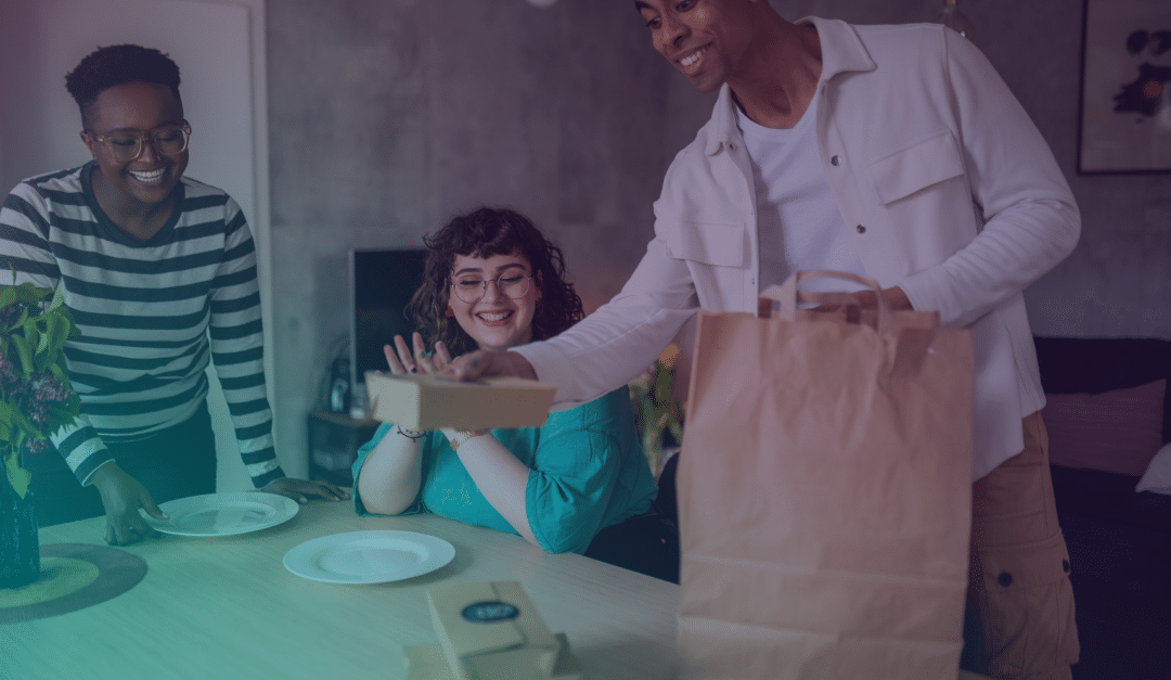 Tres personas se reúnen alrededor de una mesa con platos, sonriendo mientras una de ellas saca recipientes con comida de una bolsa de papel marrón, preparándose para comer juntos en casa.