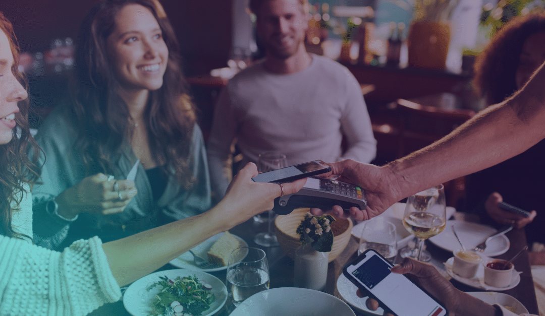 Cuatro personas están sentadas en la mesa de un restaurante con comida y bebida. Una de ellas paga la cuenta con un smartphone, mientras las demás observan y sonríen.