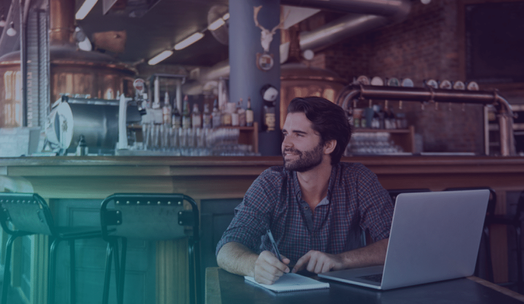Un hombre con barba está sentado en una mesa de un bar, sonriendo mientras escribe en un cuaderno con un ordenador portátil abierto delante de él. Al fondo se ven máquinas industriales de elaboración de cerveza.