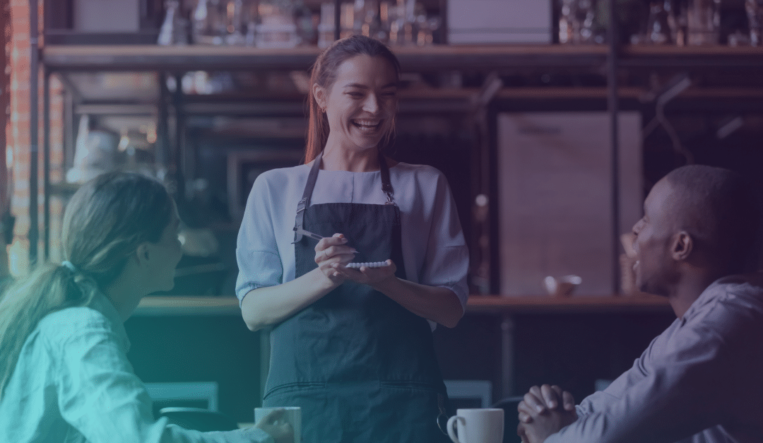 Un camarero sonriente, con delantal, está junto a una mesa, hablando con dos clientes que están sentados y sostienen tazas de café en un acogedor ambiente de cafetería.