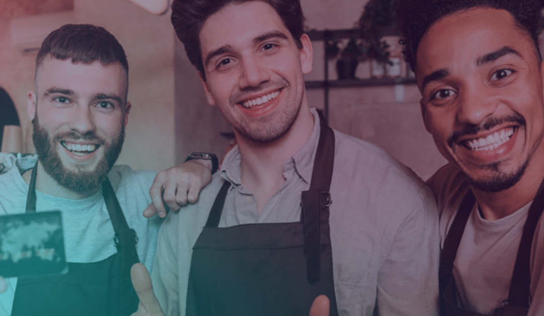 Tres hombres sonrientes, que llevan delantales, posan juntos para hacerse un selfi en lo que parece ser una cafetería o un restaurante.