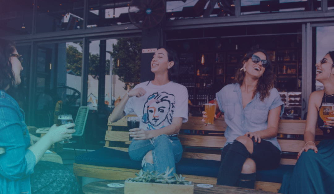 Cuatro mujeres están sentadas a la puerta de un bar, riendo y charlando mientras sostienen sus bebidas. Una de ellas lleva una camiseta con la imagen de una cara estampada. Están sentadas en bancos de madera frente a unos grandes ventanales, con plantas y vasos de cerveza sobre la mesa.