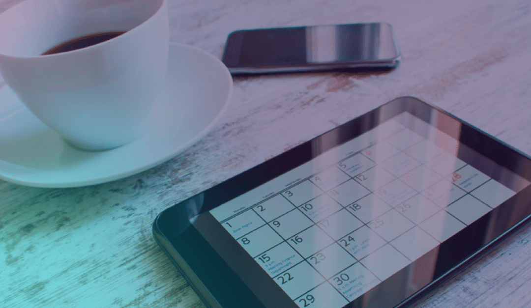 Sobre una mesa de madera, junto a una taza de café y un smartphone, hay una tableta en la que se ve un calendario.