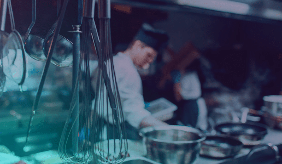 Un chef está trabajando en una cocina junto a una encimera; en primer plano se ven cuencos para mezclar y batidores colgados; la escena está ligeramente desenfocada.