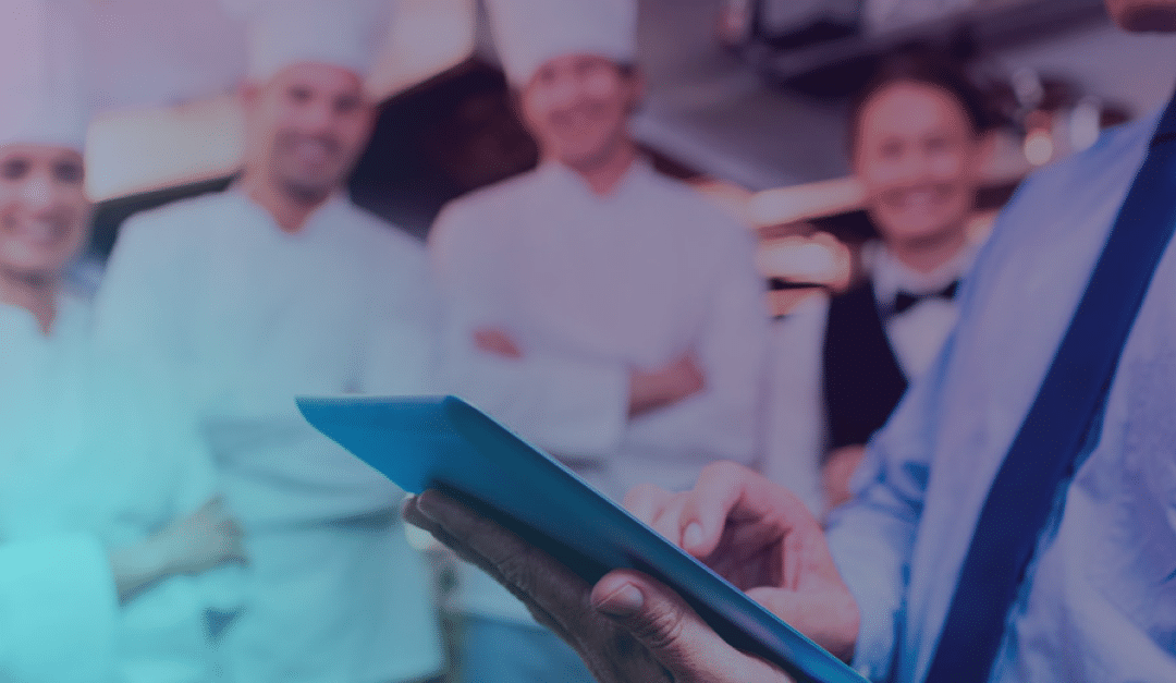 Una persona que utiliza una tableta en primer plano, con un grupo de cocineros y un camarero de pie juntos en un fondo desenfocado; todos sonríen y visten ropa de cocina profesional.