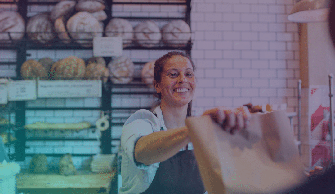Un empleado de la panadería, con una sonrisa, le entrega una bolsa de papel a un cliente, con estanterías repletas de panes variados y barras de pan al fondo.