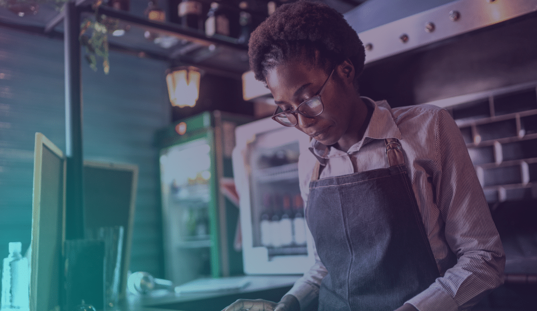 Una persona que lleva gafas y un delantal trabaja en una cocina o un restaurante, concentrada en una tarea. Al fondo se ven estanterías, una nevera con botellas y utensilios de cocina.