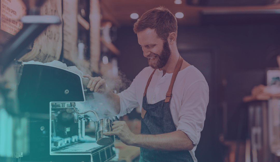 Un barista con barba y delantal sonríe mientras calienta leche con vapor en una máquina de café expreso en una acogedora cafetería.