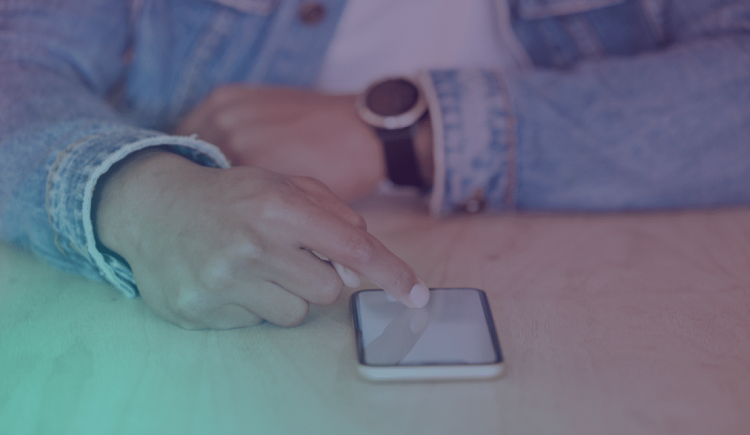 Una persona que lleva una chaqueta vaquera y un reloj está utilizando un smartphone sobre una mesa de madera, tocando la pantalla con el dedo índice.