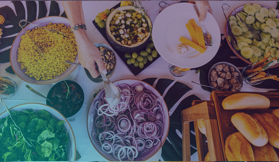 Una persona sirve comida de una colorida mesa de bufé con ensalada, cebolla cortada, maíz, pepinos, pan, queso, uvas y diversos ingredientes para acompañar en los platos.