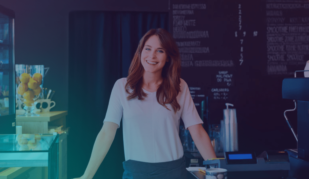 Una mujer está de pie, sonriendo, detrás del mostrador de una cafetería moderna, con una caja registradora, una vitrina de cristal y una pizarra con el menú escrito en el fondo.