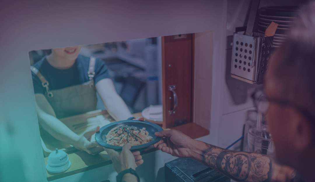 En un restaurante, una persona le pasa un plato de comida a través de una ventanilla de cocina a otra persona que tiene los brazos tatuados.