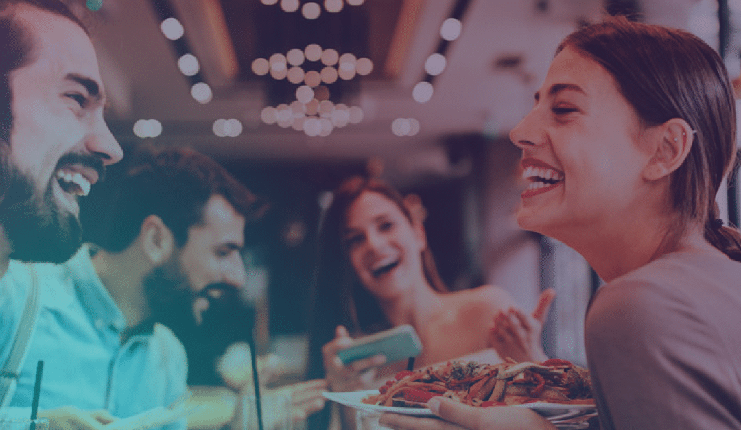 Cuatro personas están sentadas en la mesa de un restaurante, riendo y disfrutando de la comida juntas. Una de ellas sostiene un plato de pasta y otra, un smartphone. El fondo aparece ligeramente desenfocado, con cálidas luces decorativas.