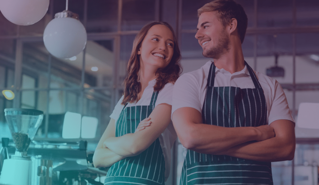 Dos personas sonrientes, con delantales a rayas, están de espaldas la una a la otra, con los brazos cruzados, en una cafetería moderna, mirándose entre sí.
