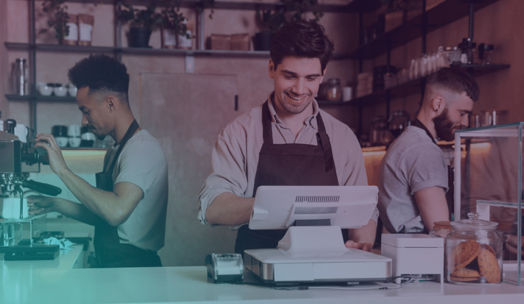 Tres empleados de la cafetería detrás del mostrador: uno prepara café, otro sonríe mientras maneja la caja registradora y el tercero prepara la comida. Se ven tarros de galletas y utensilios de cocina.