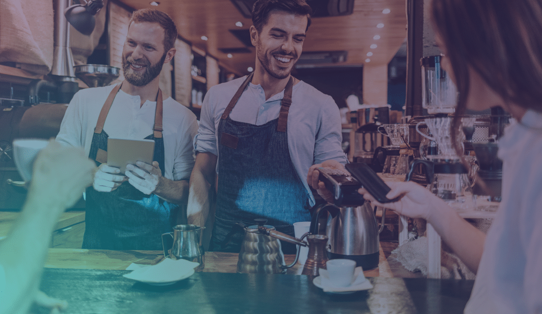 Dos baristas, con delantal, sonríen detrás de la barra de una cafetería mientras un cliente paga con un smartphone. En la barra de madera se ven tazas y utensilios de café.