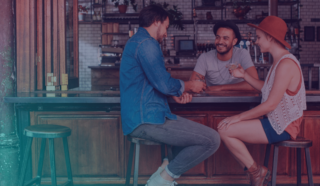 Tres personas están sentadas en un bar, sonriendo y charlando. Dos de ellas están sentadas en taburetes, mientras que el camarero está de pie detrás de la barra. El ambiente es informal, con una decoración de madera y las bebidas en la mano.