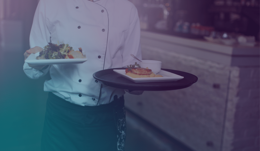 Un camarero, vestido con una chaqueta blanca de chef, lleva dos platos con comida, uno en cada mano, en el interior de un restaurante con un fondo desenfocado.