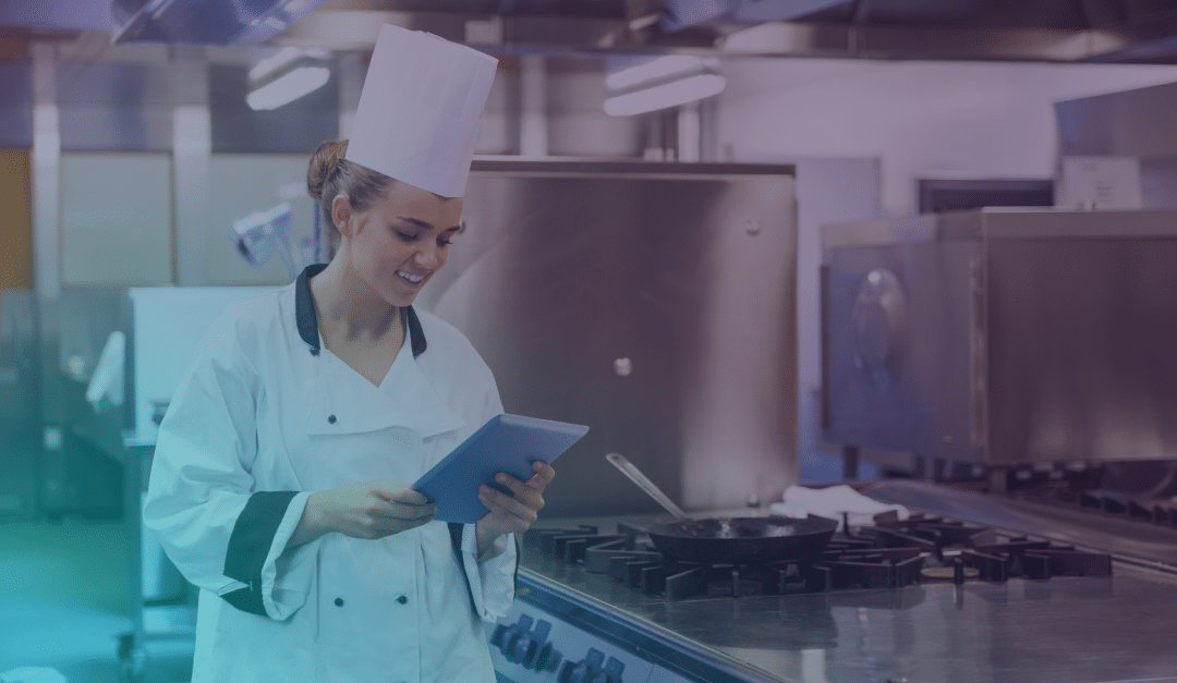 Un chef, vestido con un uniforme blanco y un gorro, está de pie en una cocina industrial, sonriendo mientras mira una tableta. Cerca de allí, hay una sartén sobre la placa de cocina.