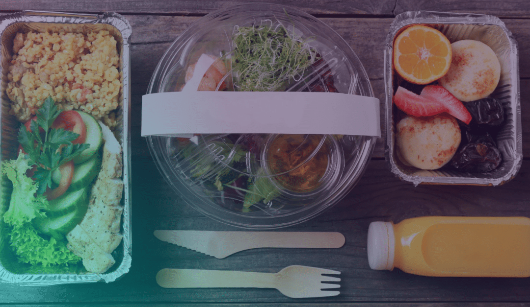 Una comida para llevar con arroz, ensalada, fruta, postre y una botella de zumo sobre una mesa de madera. Delante de los recipientes hay un tenedor y un cuchillo de madera.