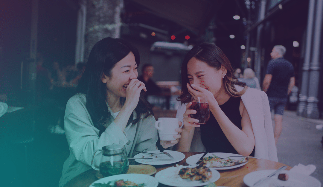 Dos mujeres están sentadas en la terraza de una cafetería, riendo juntas mientras disfrutan de una bebida y algo de comer. Al fondo se ven otros comensales y un peatón.