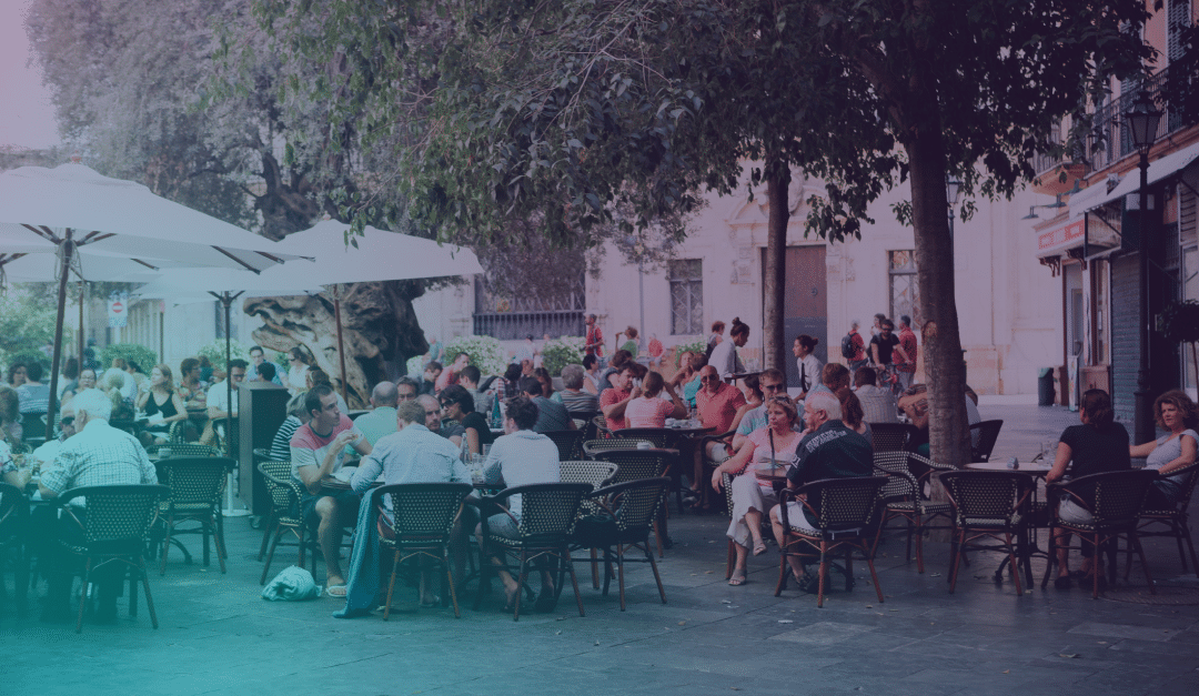 La gente se sienta en las mesas de una cafetería al aire libre, bajo las sombrillas y los árboles de una plaza animada, charlando y comiendo. El ambiente es sombreado y relajado, con edificios y vegetación al fondo.
