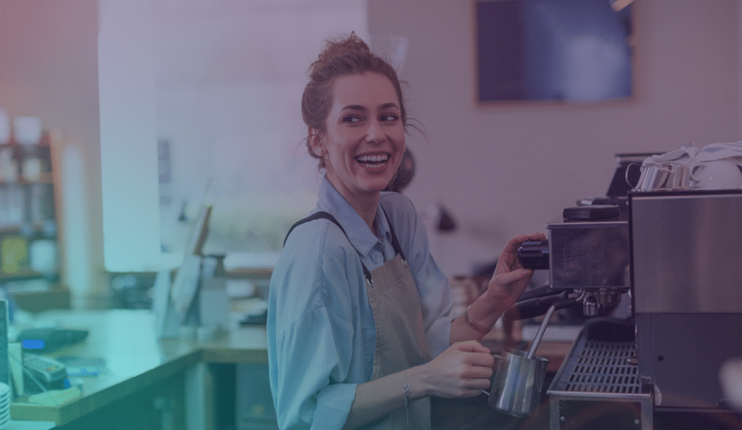 Un barista sonriente, con delantal, está de pie detrás de una cafetera, calentando leche con vapor en una cafetería. Al fondo se ven estanterías y la decoración difuminada de la cafetería.