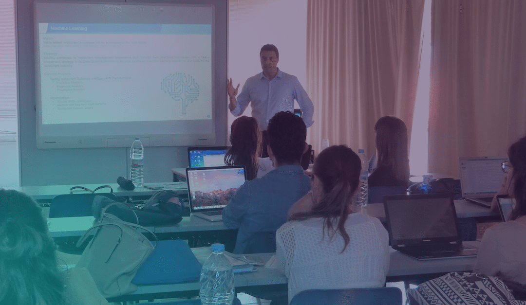 Un hombre está de pie frente a un aula impartiendo una presentación sobre aprendizaje automático, mientras los alumnos, sentados en sus pupitres, utilizan sus ordenadores portátiles y escuchan con atención.