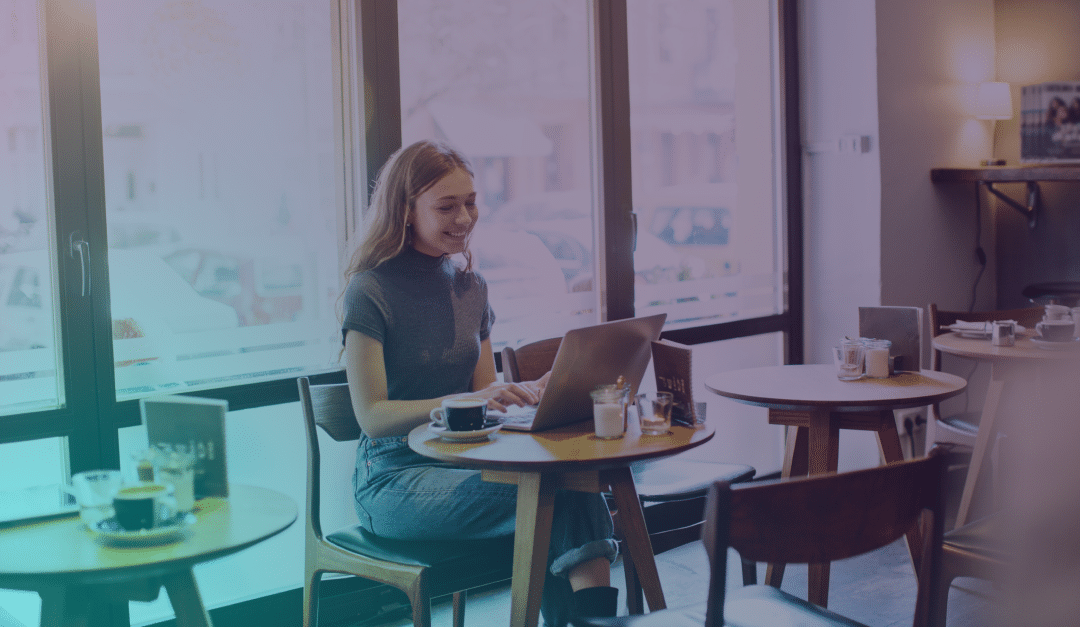 Una mujer está sentada en una mesa de una cafetería, sonriendo a su ordenador portátil. Tiene una taza de café y un bloc de notas a su lado, con grandes ventanales al fondo que dejan entrar la luz natural.