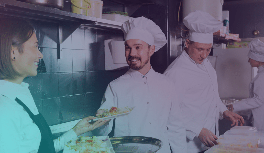 Tres cocineros con uniformes blancos se encuentran en una cocina industrial. Uno de ellos sonríe y le entrega un plato con comida a una mujer, mientras otro prepara platos al fondo.